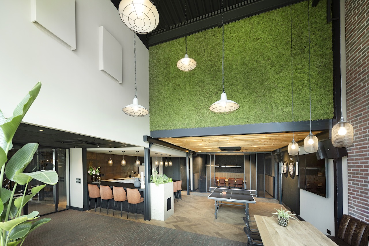 Ruimtelijk overzicht van de multifunctionele kantine van de Van der Werff Groep, met een grote groene moswand, een bar met barkrukken, een tafeltennistafel en diverse industriële hanglampen.