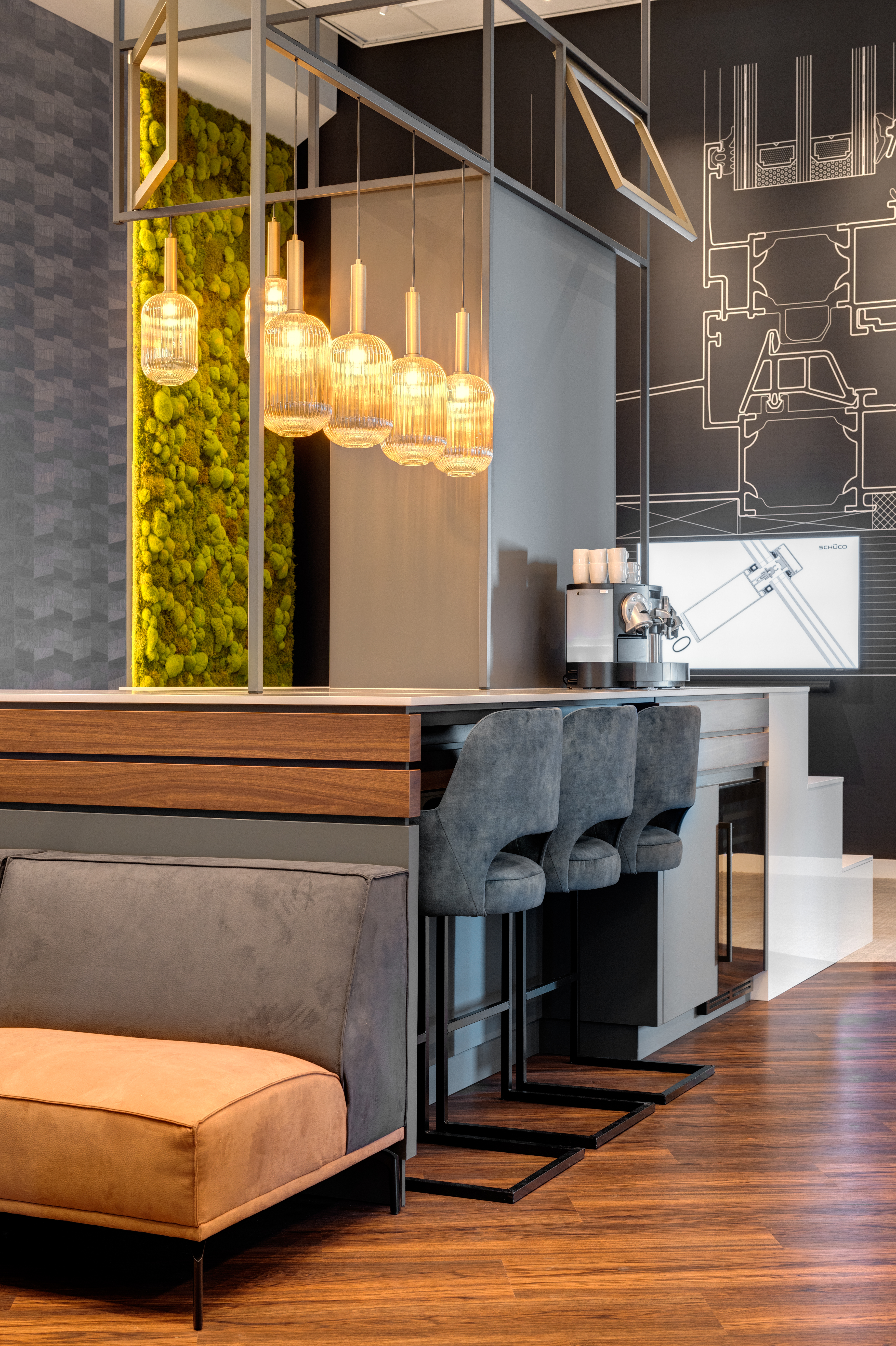 Een modern interieur van een luxe zithoek en bar-ruimte. Op de voorgrond een cognac-kleurige bank met een donkergrijze rugleuning, geplaatst tegen een donkergrijze gevelbeplating en een houten lambrisering. De bar-ruimte erachter heeft een donker houten aanrechtblad met een roestvrijstalen kraan, een espressomachine. Drie donkergrijze barkrukken staan aan de bar. Boven de bar hangen een reeks opvallende goudkleurige, geribbelde glazen hanglampen aan een modern, open, metalen plafondframe.