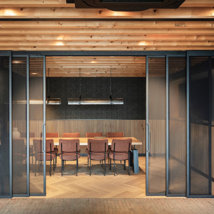 Van der Werff Groep vierkant Industrieel-chique vergaderruimte van de Van der Werff Groep, gezien door geopende zwarte metalen gaasdeuren, met roze gecapitonneerde stoelen, een houten tafel en akoestische lattenwanden.